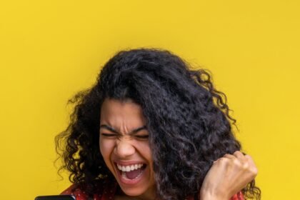 Beautiful dark skinned girl holding mobile phone and making winner's gesture clenching her fist, screaming yes celebrating success, expressing joy about victory in online lottery.